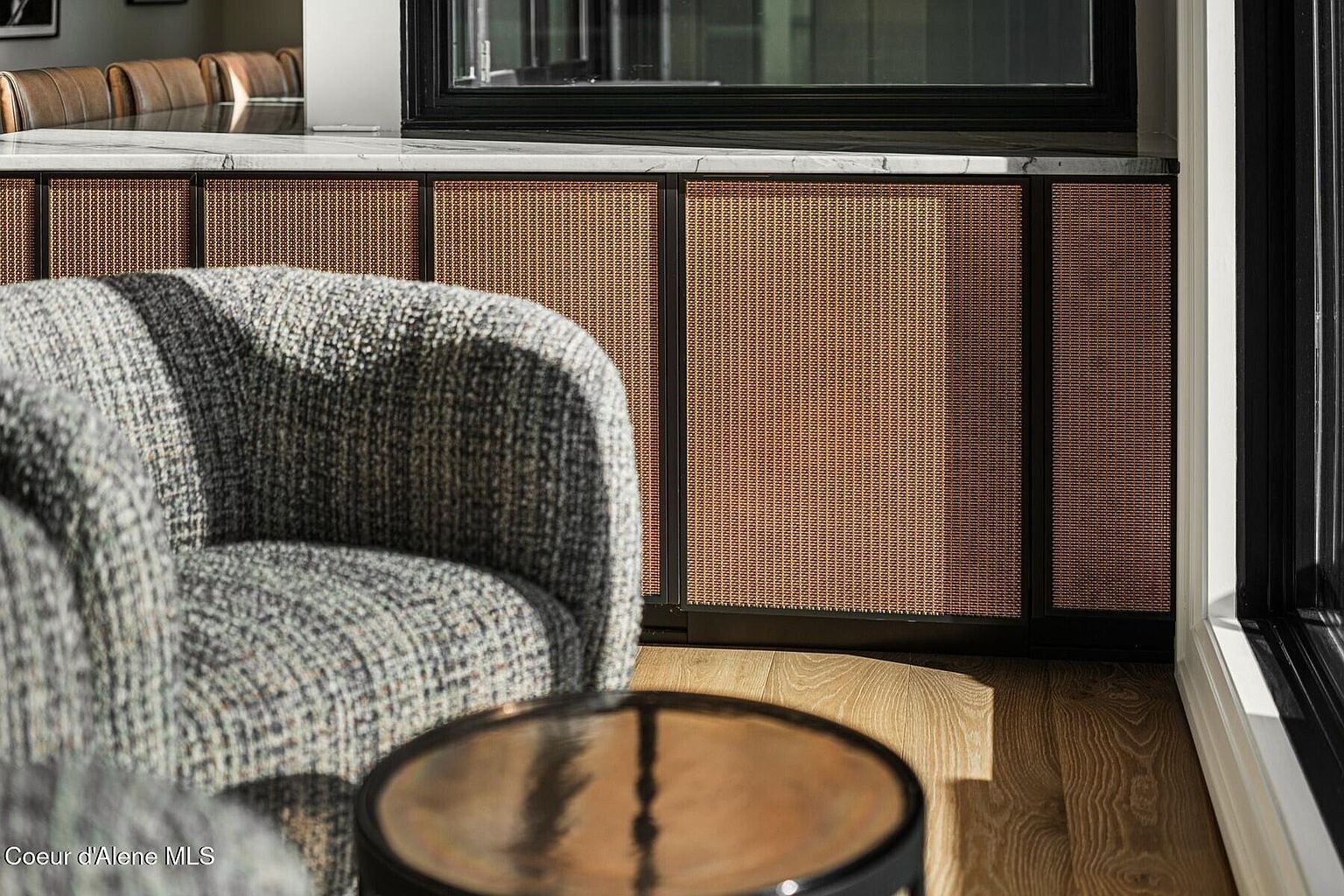 This interior shot showcases a cozy living room setting with a focus on texture and design. A patterned armchair sits beside a small, reflective side table, with a backdrop of woven-panel cabinetry beneath a marble countertop. The room is bathed in natural light, highlighting the warm wood flooring and creating an inviting atmosphere.