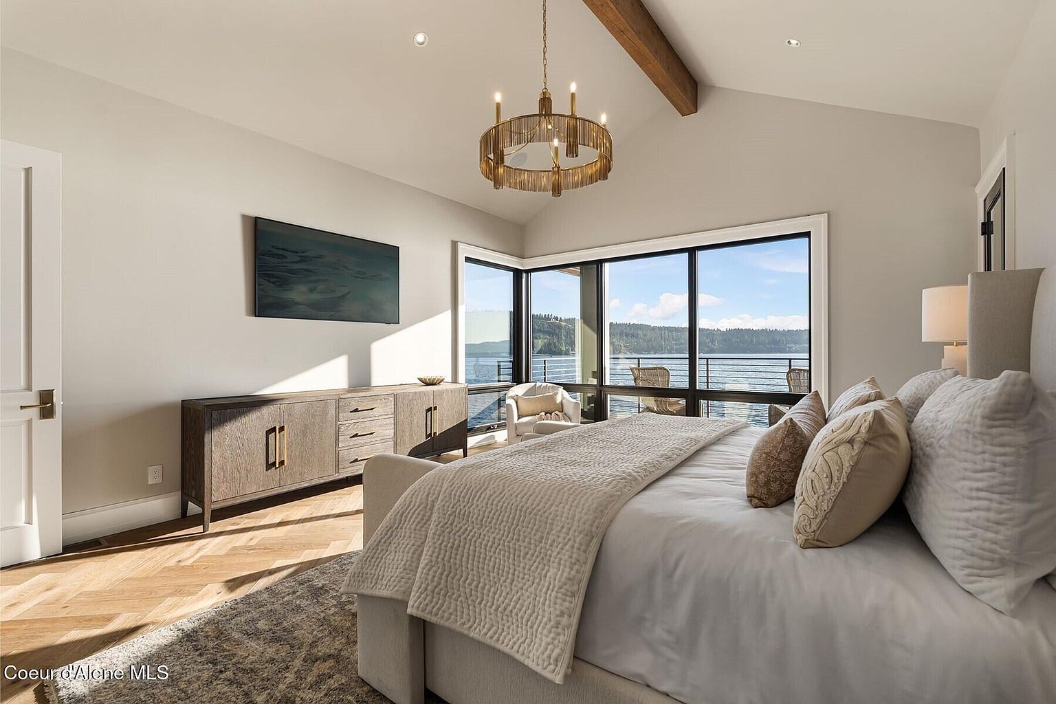 This is a luxurious primary bedroom featuring a large bed with plush pillows and a textured throw. A wooden dresser sits against the wall beneath a modern art piece, while a statement chandelier hangs from the vaulted ceiling with a wooden beam. Large windows offer stunning views of the water, creating a serene and inviting atmosphere.