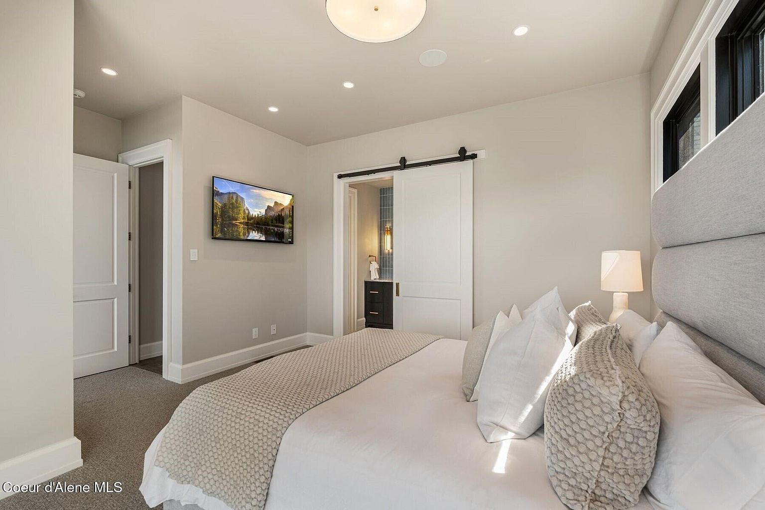 This is a primary bedroom featuring a large bed with a padded headboard, white linens, and decorative pillows. A sliding barn door leads to an en-suite bathroom. A television is mounted on the wall, and recessed lighting illuminates the space, creating a serene and inviting atmosphere.