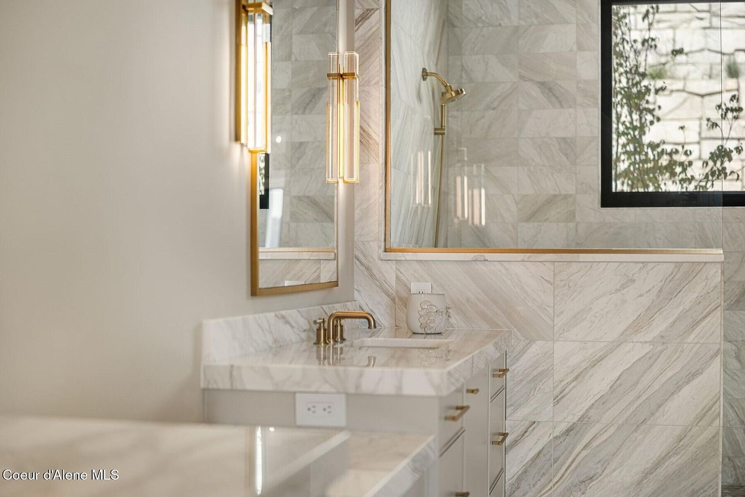 This is a well-lit primary bathroom featuring marble-like tile walls and countertops, gold fixtures, and modern lighting. The vanity area includes a sink, storage cabinets with gold hardware, and a mirror reflecting the shower area. A window provides natural light and a view of the outdoors, enhancing the luxurious and spa-like atmosphere.
