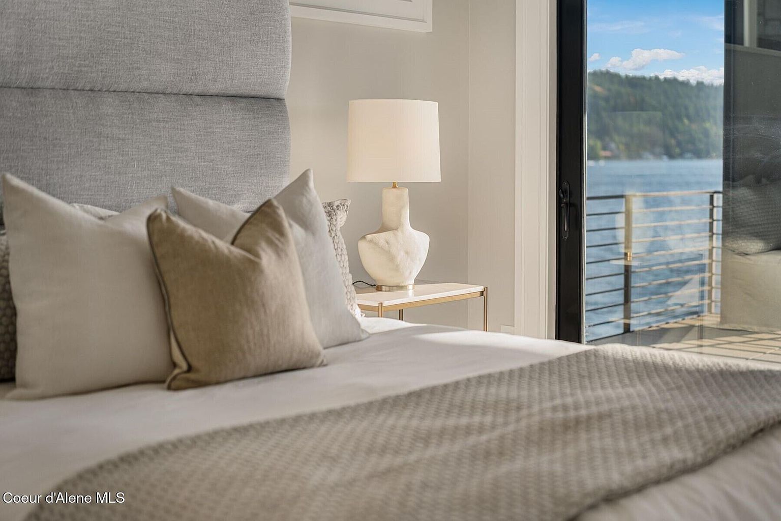 This is a serene primary bedroom featuring a plush bed with neutral-toned pillows and a textured throw. A modern lamp sits on a side table, and a sliding glass door offers a view of a lake or ocean. The room exudes a calming and luxurious atmosphere.