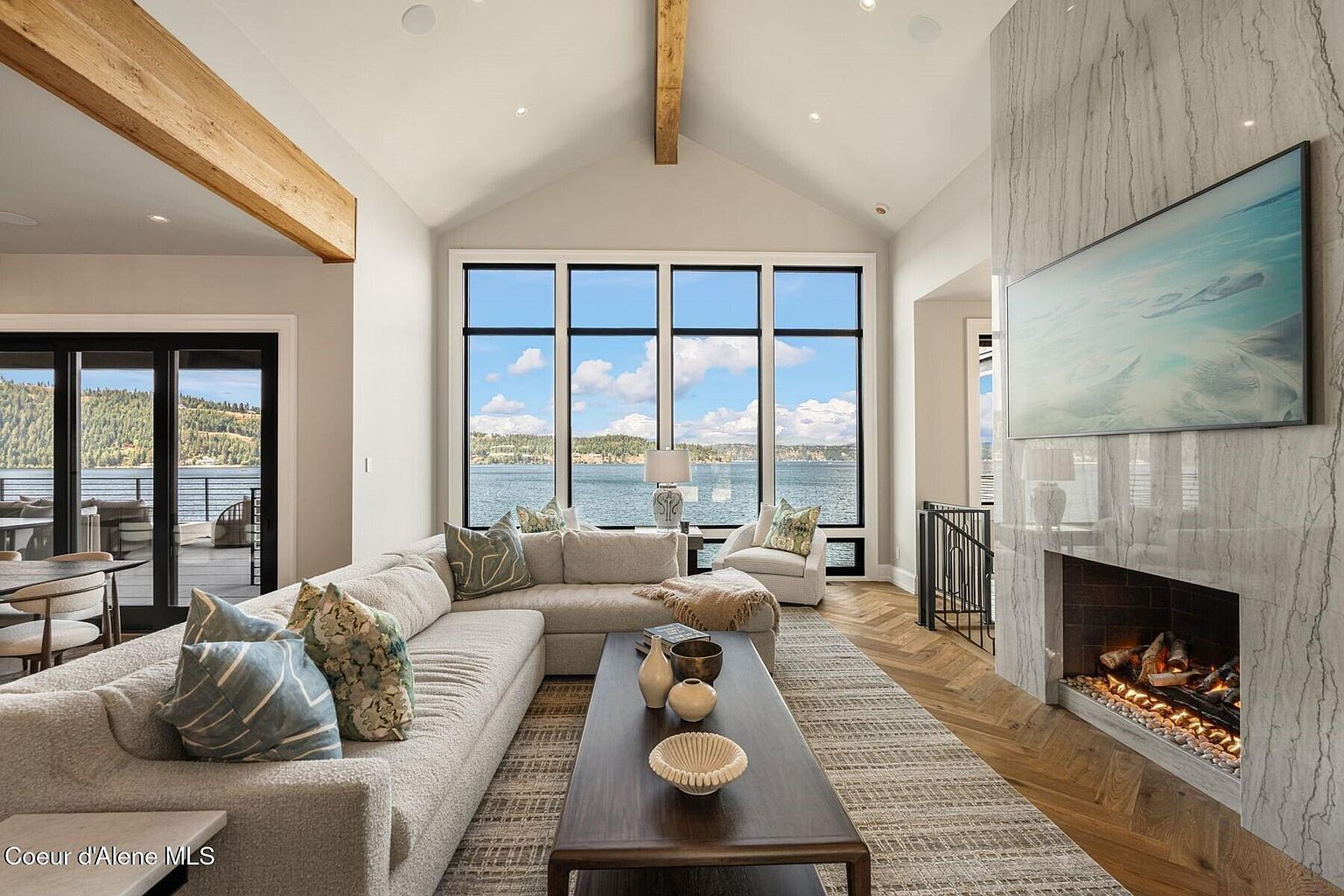 This is a bright and airy living room featuring a large sectional sofa, a dark wood coffee table, and a striking fireplace with a marble surround. The room boasts expansive windows offering stunning lake views, complemented by exposed wooden beams that add a touch of rustic charm. The open layout and neutral color palette create a welcoming and luxurious atmosphere.