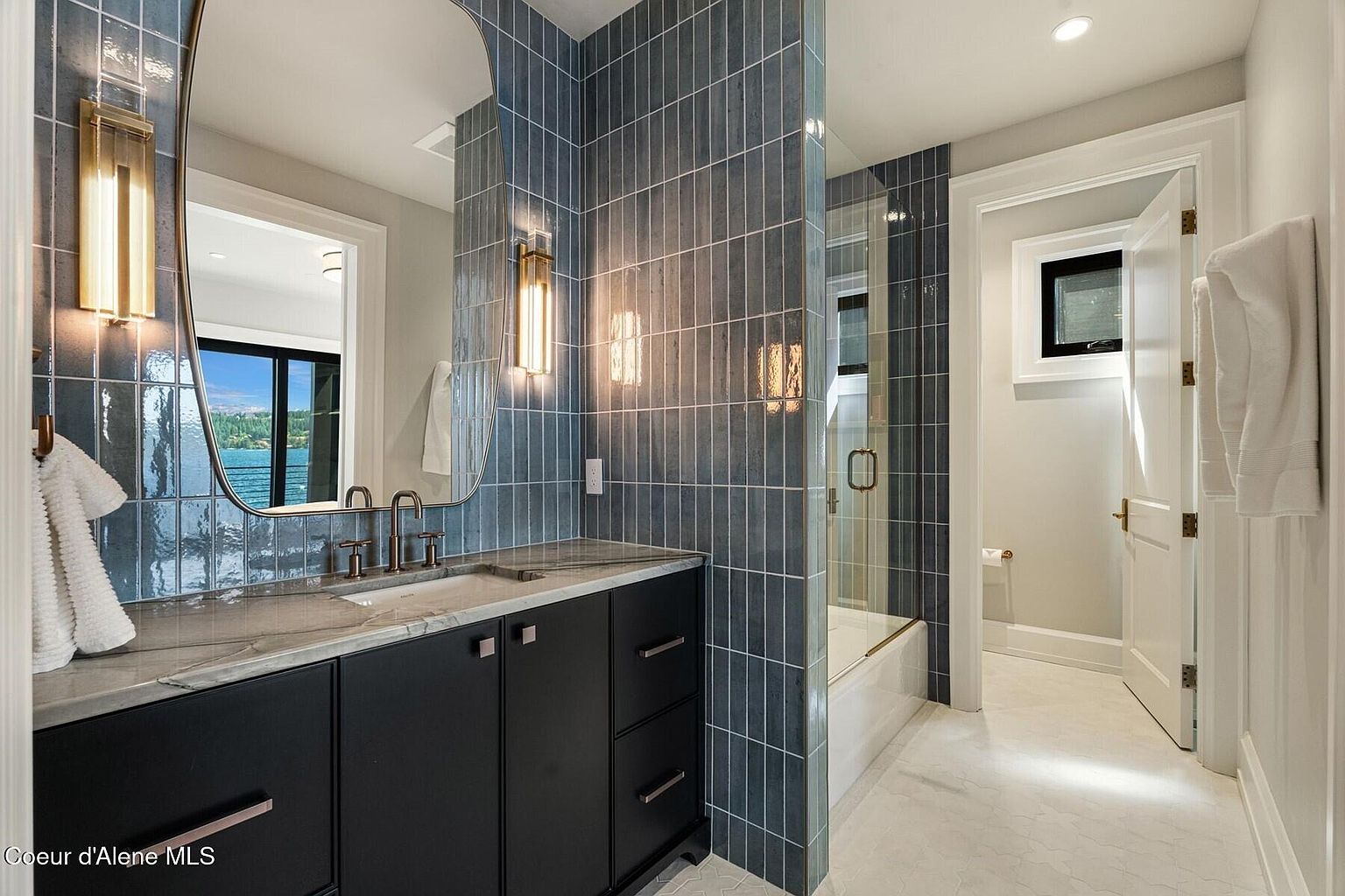 This is a well-designed bathroom featuring blue vertical tiles, a dark vanity with a marble countertop, and modern fixtures. The space includes a glass-enclosed shower and a separate toilet room, all contributing to a clean and contemporary aesthetic. The mirror reflects a view of the outside, adding depth and interest to the room.