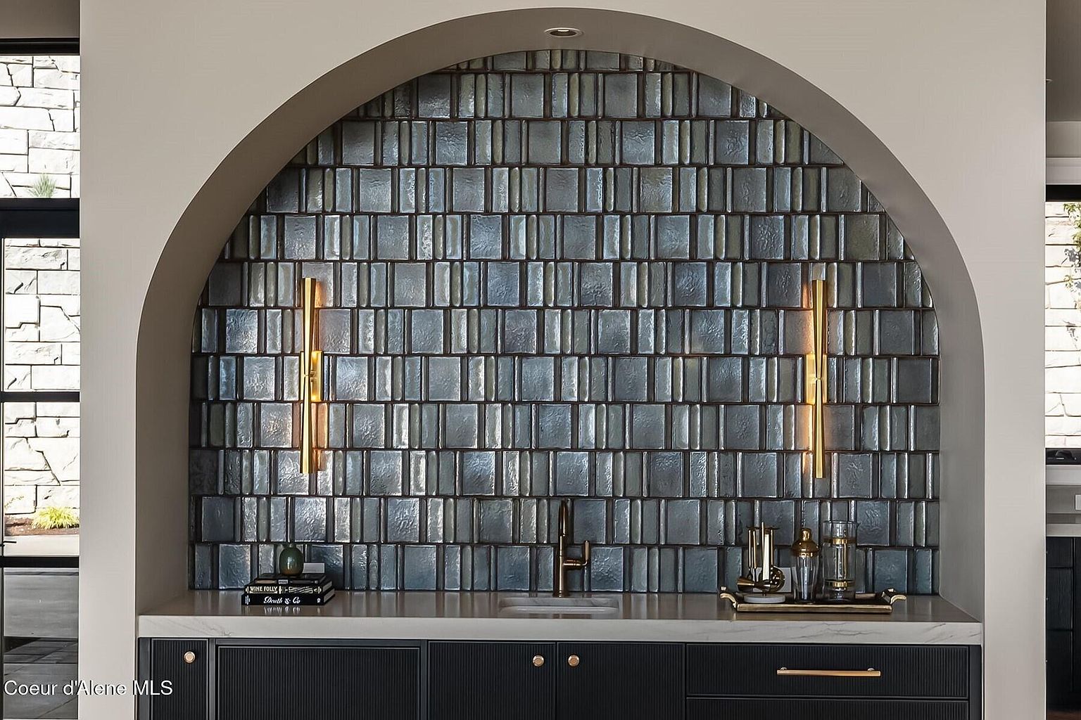 This image showcases a stylish bar area within a kitchen, featuring a striking arched alcove with textured blue-gray tile backsplash. The countertop is light-colored, contrasting with the dark cabinetry below, which is accented with gold hardware. Two vertical sconces flank the backsplash, adding warm lighting to the scene.