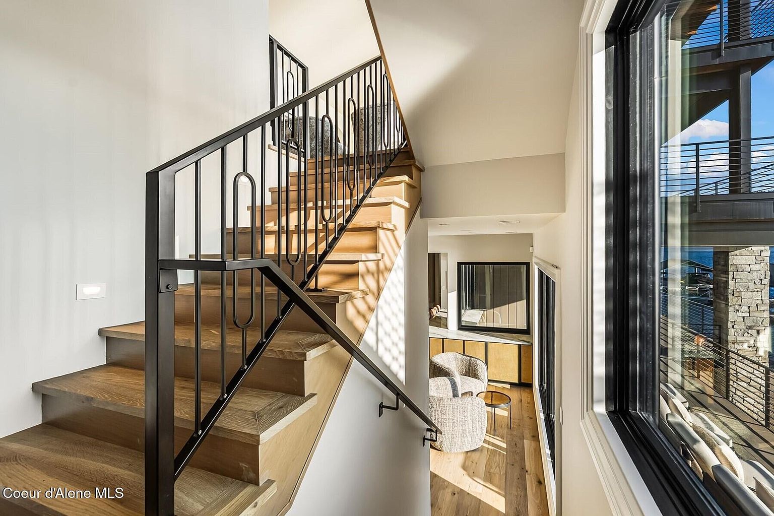 This interior shot showcases a modern hallway with wooden stairs and a black metal railing. Natural light floods the space through a large window, highlighting the light wood flooring and neutral wall colors. The hallway leads to a sitting area with stylish chairs, creating an inviting and sophisticated atmosphere.