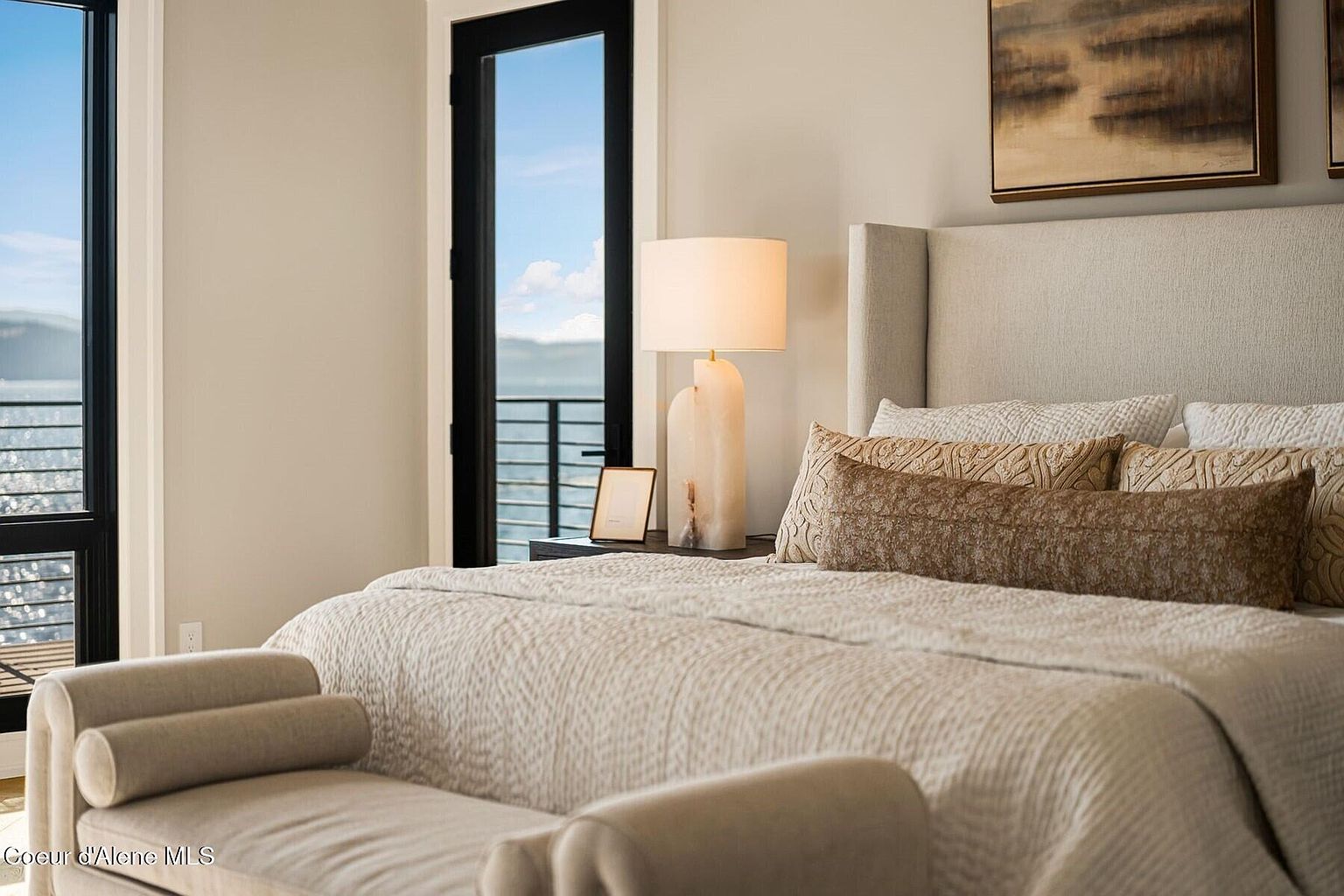 This is a serene primary bedroom featuring a large bed with a neutral-toned quilted coverlet and decorative pillows. A modern lamp sits on the nightstand next to a framed picture. The room has a calming atmosphere with a view of the lake through a black-framed door, and a bench sits at the foot of the bed.