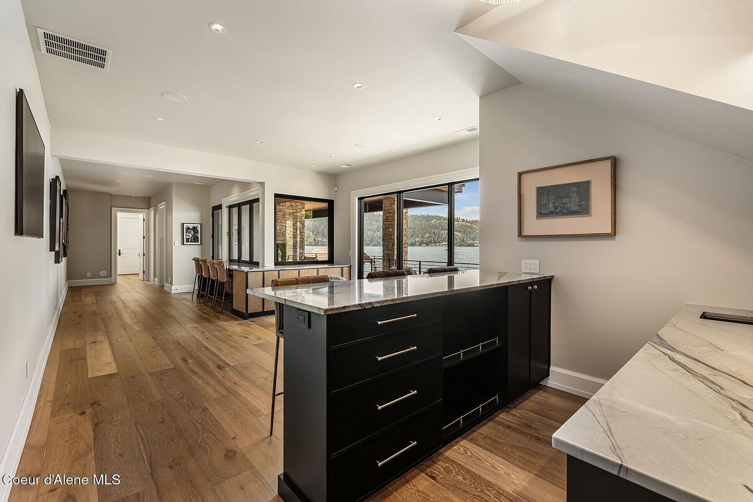 This interior shot showcases a modern kitchen with hardwood flooring and a black kitchen island topped with marble. Large windows offer a view of the lake, enhancing the natural light in the space. The kitchen seamlessly blends functionality with a stylish design, perfect for entertaining and everyday living.