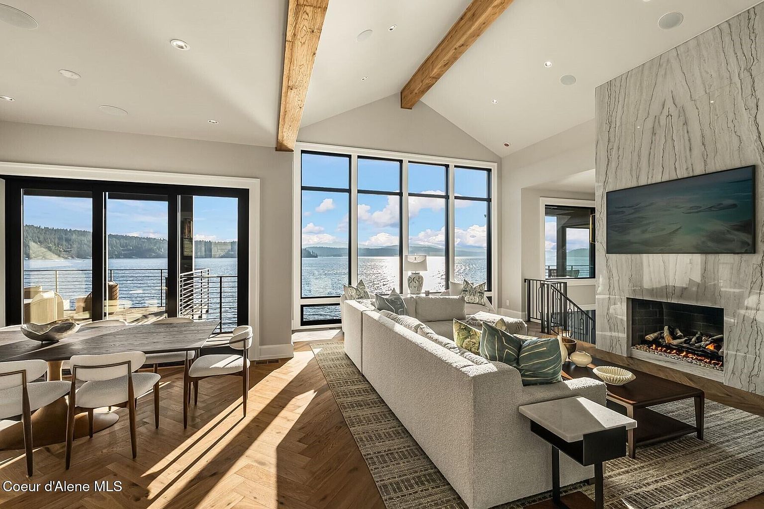 This is an interior shot of a luxurious living room with a stunning waterfront view. The room features a large sectional sofa, a modern fireplace with a marble surround and a mounted television, and exposed wooden beams on the vaulted ceiling. The open floor plan includes a dining area with a modern table and chairs, all complemented by the natural light flooding in through the expansive windows.