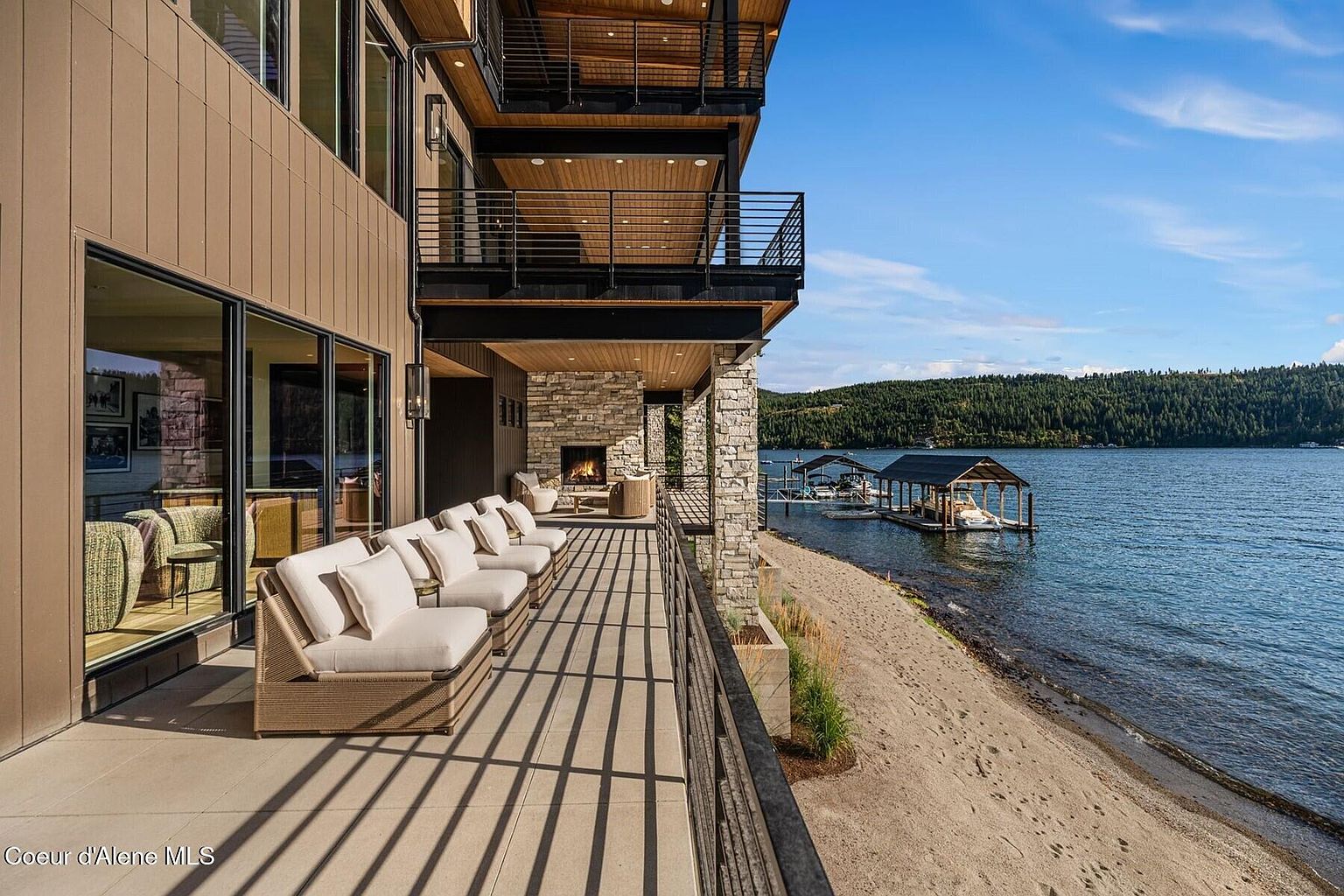 This image showcases a luxurious outdoor patio/deck/balcony area of a waterfront property. The space features comfortable lounge seating, a stone fireplace, and stunning views of the lake and surrounding landscape. The design emphasizes relaxation and outdoor living, making it an ideal selling point for potential buyers.