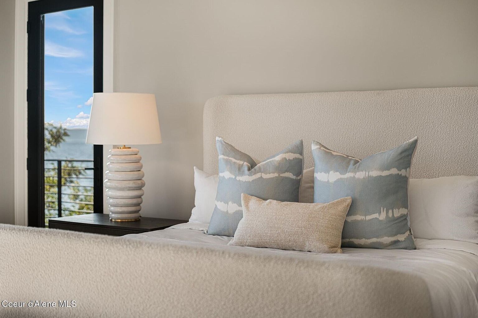 This is a serene primary bedroom featuring a neutral color palette. The bed is adorned with decorative pillows in varying textures and patterns, complemented by a textured headboard. A stylish lamp sits on the bedside table, and a glimpse of an outdoor view is visible through a nearby window, creating a calming and inviting atmosphere.