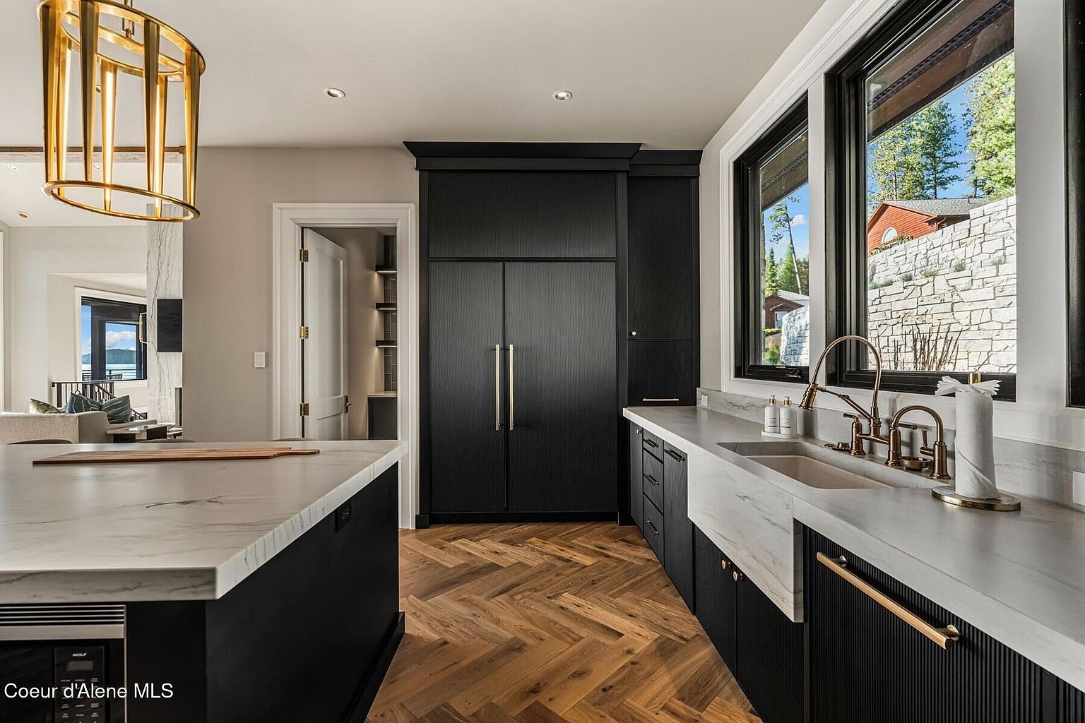 This is a modern kitchen with black cabinetry and gold hardware, featuring a large island with a white marble countertop. The flooring is a herringbone pattern wood, and there are stainless steel appliances. The kitchen also has a large window overlooking the exterior, and a gold chandelier hangs above the island.