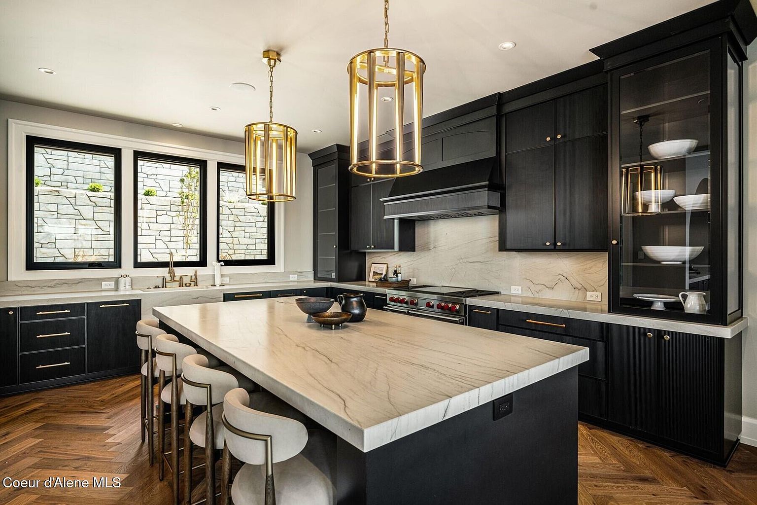 This is a modern kitchen featuring black cabinetry with gold hardware and a large island with a light-colored countertop. Two gold pendant lights hang above the island, and a window provides natural light. The kitchen also includes stainless steel appliances and a marble backsplash, creating a luxurious and stylish space.