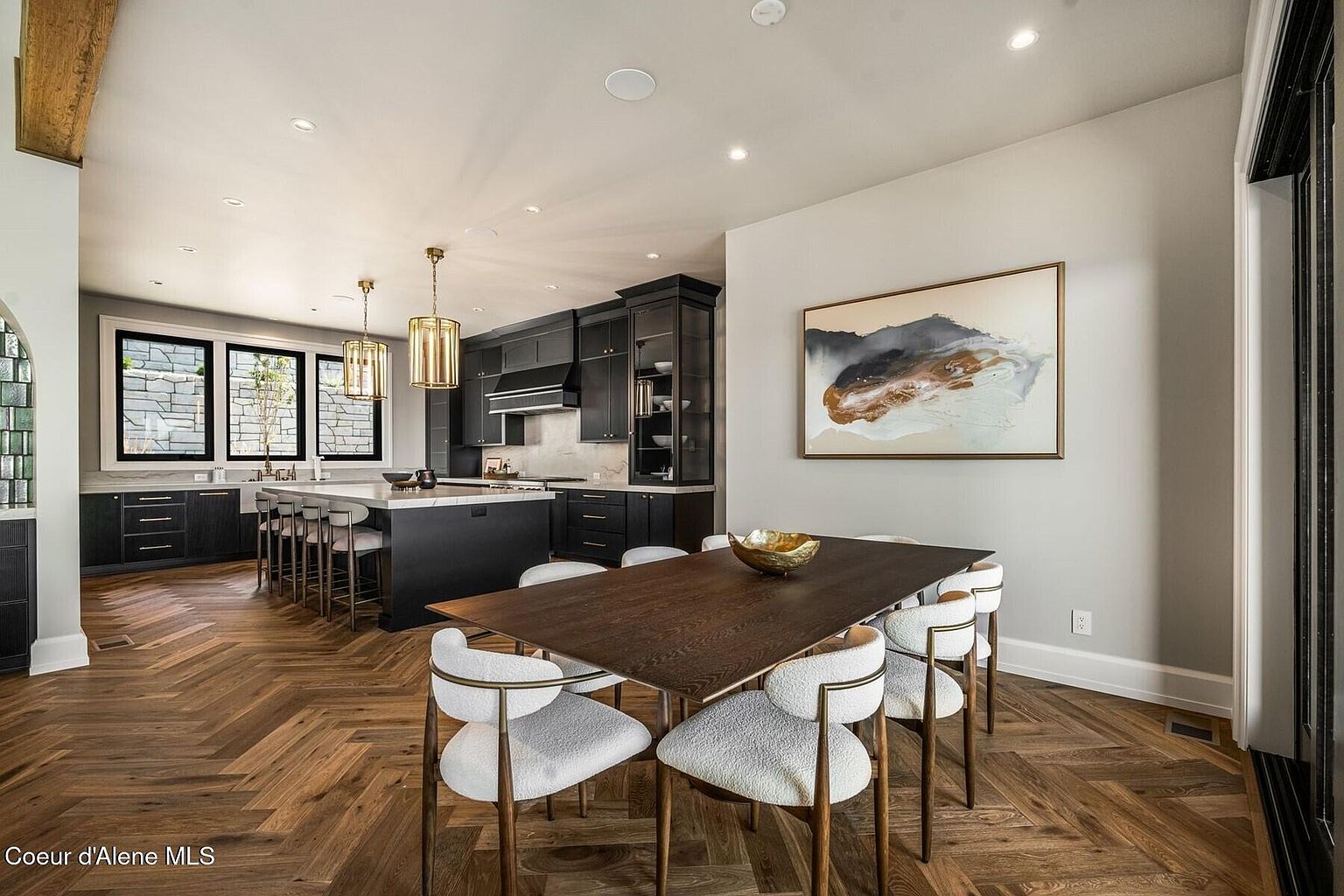 This interior shot showcases a dining area adjacent to a modern kitchen. The dining room features a dark wood table surrounded by stylish chairs with white upholstery and gold frames. An abstract painting hangs on the wall, and the herringbone wood flooring adds a touch of elegance.