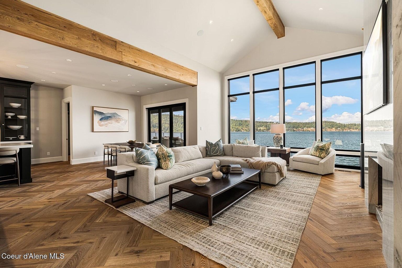 This is a spacious living room featuring a large sectional sofa, a coffee table, and an area rug. The room has large windows that offer a view of the lake. The room also has wooden beams on the ceiling and hardwood floors, which add warmth and character to the space.