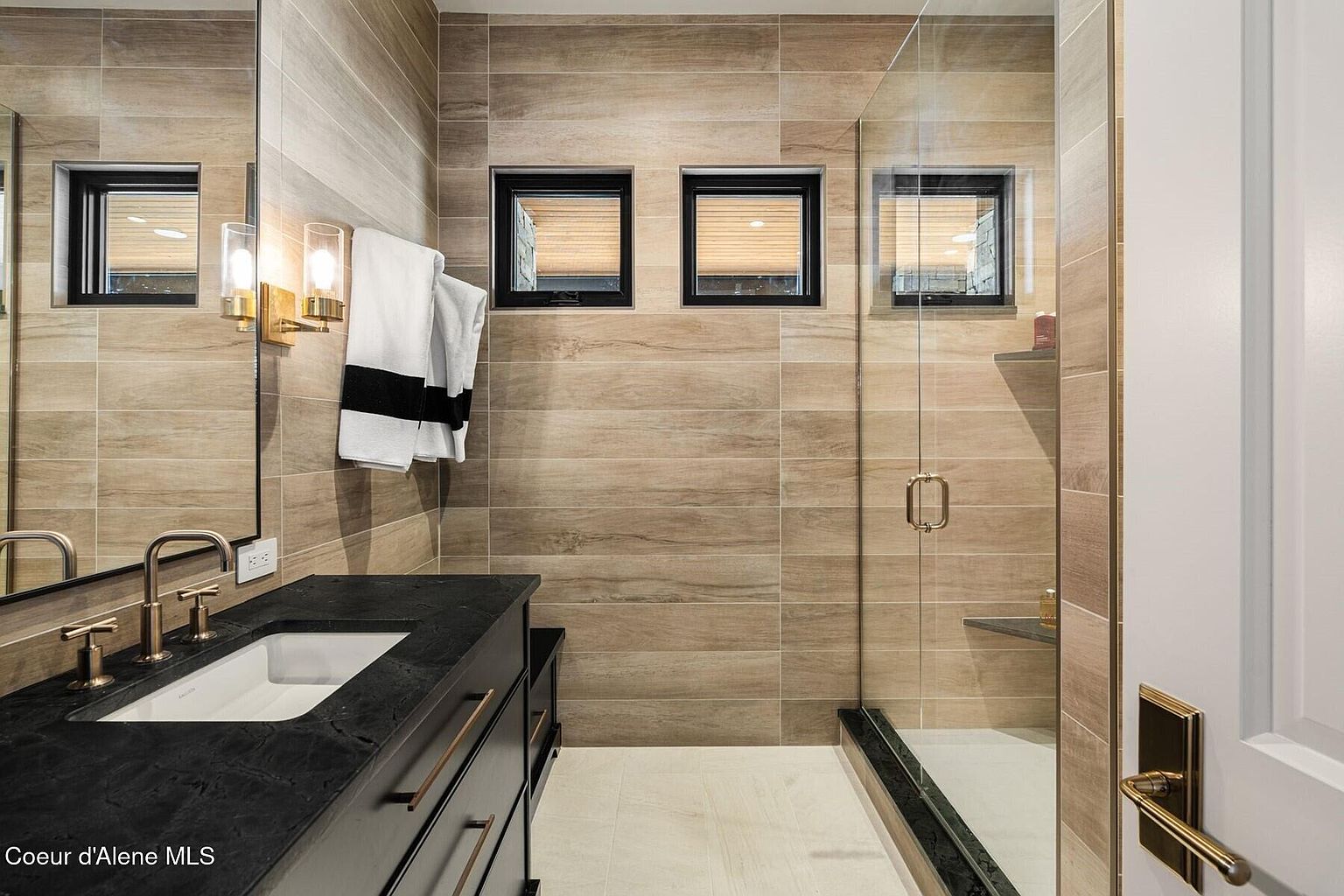 This is a modern bathroom featuring a black countertop vanity with a white sink and gold fixtures. The walls are covered in horizontal wood-look tiles, complemented by three small windows above the shower area. A glass-enclosed shower with gold hardware adds to the contemporary design, while a white door with gold hardware is visible on the right.