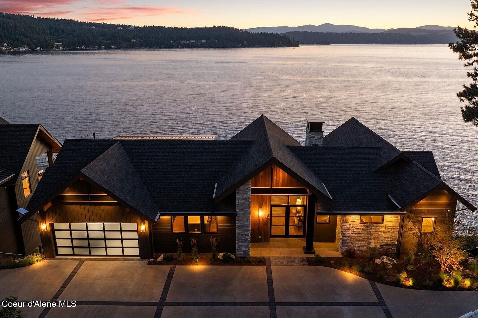This is a front exterior view of a luxurious waterfront home at dusk. The house features a dark roof, a mix of wood and stone facade, and a well-lit driveway leading to a garage. The large windows and entryway are illuminated, creating a warm and inviting atmosphere, with a serene lake and mountain backdrop adding to the property's appeal.