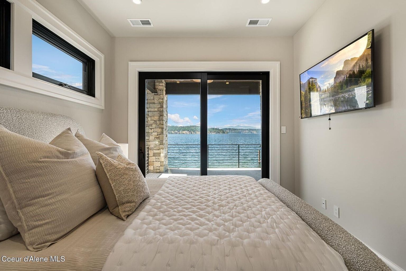 This is a primary bedroom featuring a large bed with neutral-toned bedding and pillows. A sliding glass door provides access to a balcony with a view of the water. A wall-mounted television is present, and the room is painted in a neutral color scheme, creating a serene and inviting atmosphere.