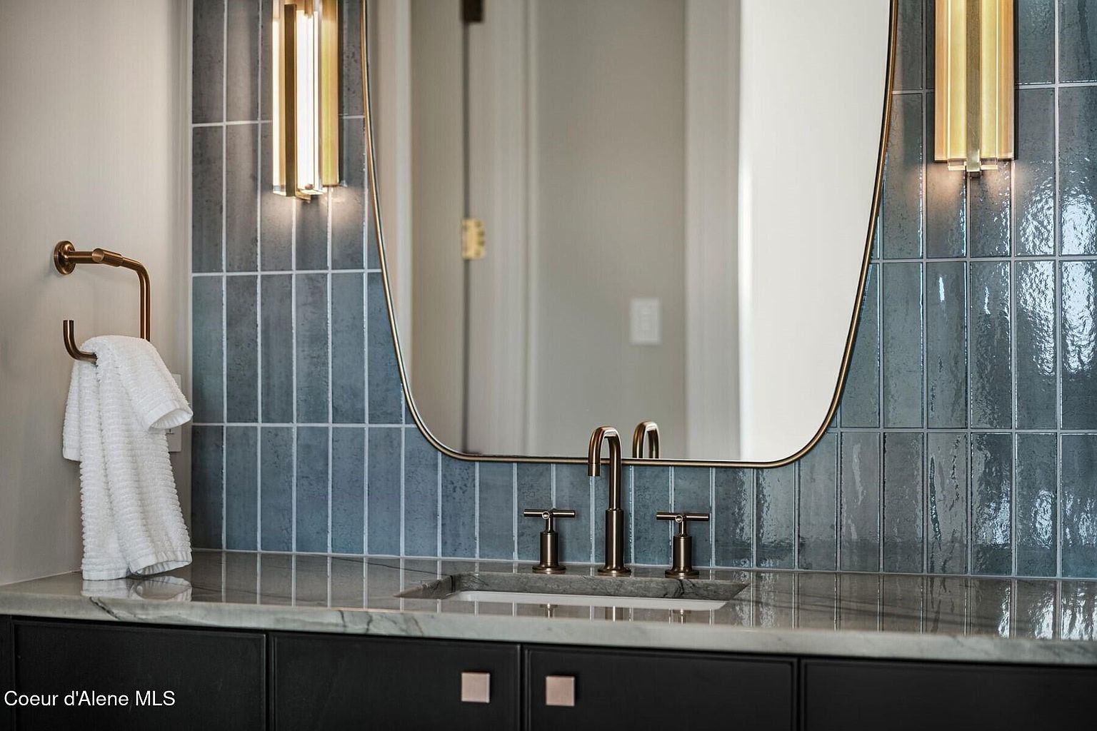 This is a detailed shot of a bathroom vanity area, showcasing a modern design with blue-gray vertical tiles and gold accents. The vanity features a dark cabinet with square hardware, a light-colored countertop, and a bronze faucet set. A decorative mirror and sconces add to the sophisticated aesthetic.