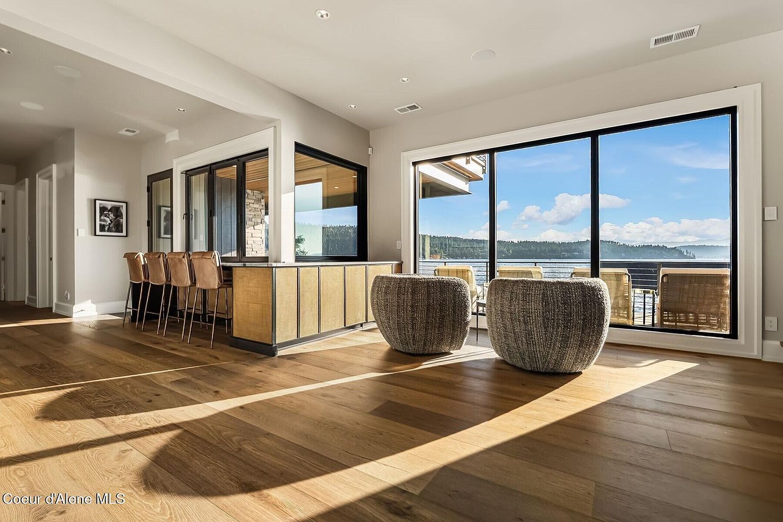 This interior shot showcases a bright and airy living room with hardwood floors and large windows offering a scenic view of the water. The room features modern furniture, including two woven chairs, and a bar area with stools, creating a stylish and inviting space. The natural light enhances the warmth and elegance of the room.