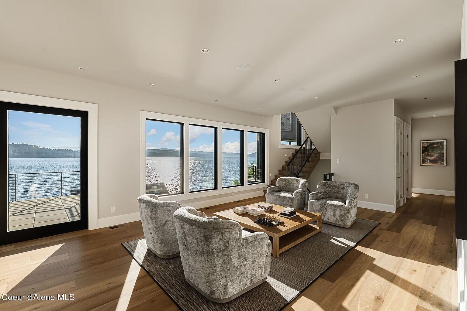 This is an interior shot of a living room with a stunning waterfront view. The room features hardwood floors, a neutral color palette, and four plush armchairs arranged around a wooden coffee table on a dark rug. Large windows frame the lake, and a staircase is visible in the background, creating a bright and inviting space.