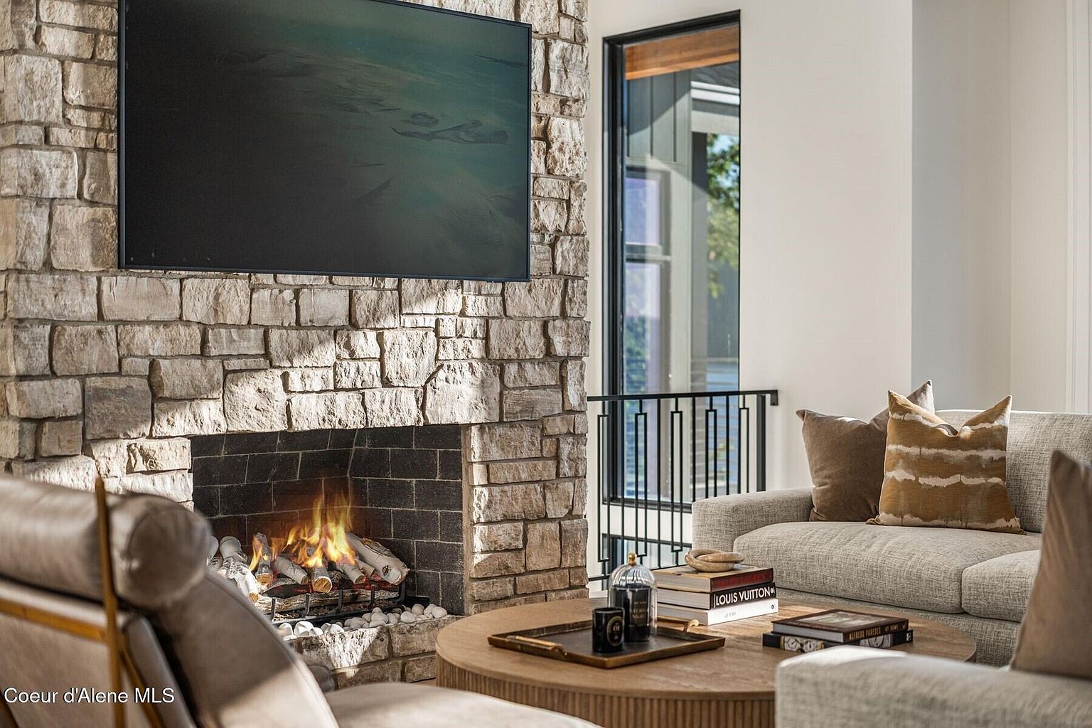 This image showcases a cozy living room with a prominent stone fireplace and a mounted television above it. The room features a comfortable-looking sofa with decorative pillows, a round wooden coffee table adorned with books and decorative items, and a glimpse of a modern window offering natural light. The overall impression is one of warmth and sophisticated comfort.