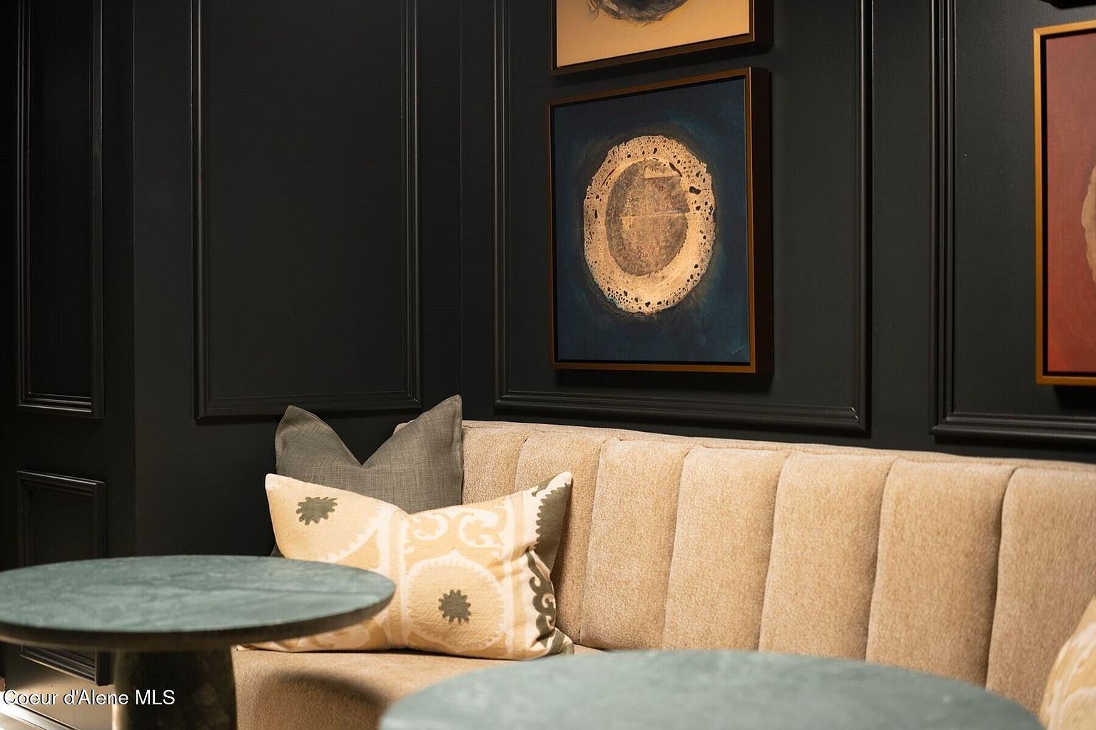 This interior shot showcases a stylish living room area with dark paneled walls adorned with framed artwork. A comfortable, tufted beige banquette seating area is accented with decorative pillows, and round green tables provide a functional surface. The overall aesthetic is sophisticated and inviting, suggesting a high-end residential space.