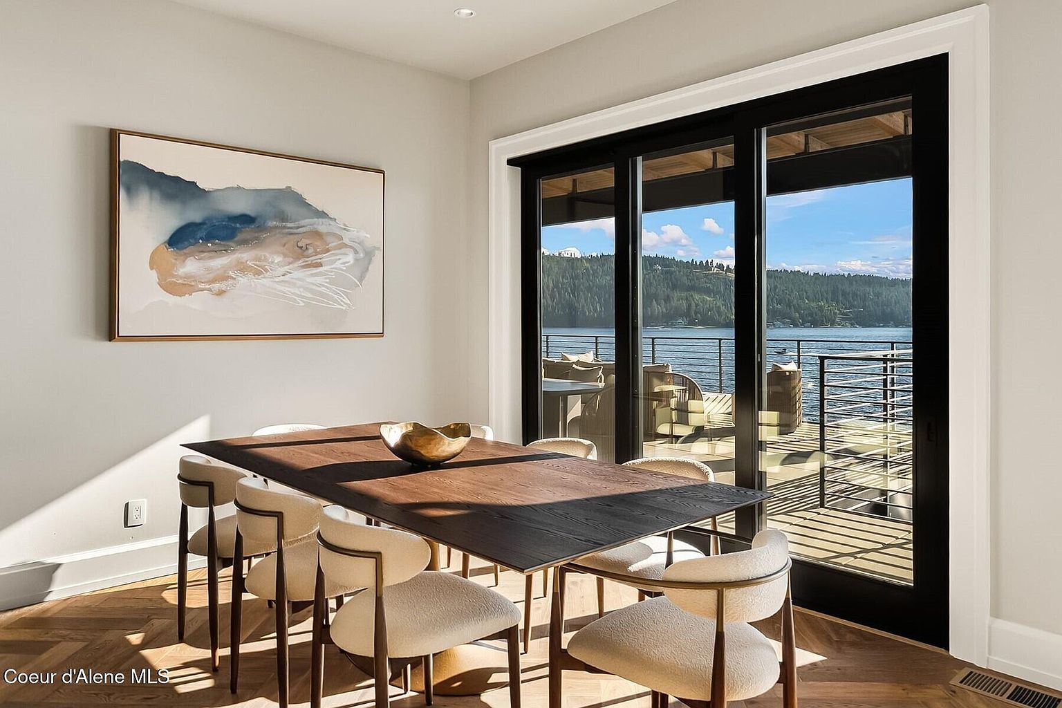 This is a dining room featuring a dark wood table surrounded by modern chairs with light-colored upholstery. A contemporary art piece hangs on the wall, and a large sliding glass door provides access to an outdoor deck with a view of a lake. The room is well-lit with natural light, creating a bright and inviting atmosphere.