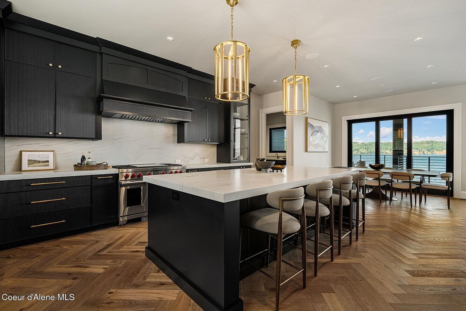 This is a beautifully designed kitchen featuring dark cabinetry with gold hardware, a large island with seating, and herringbone wood floors. The kitchen is well-lit with pendant lighting and natural light from the adjacent dining area with views of the water. The overall impression is luxurious and modern.
