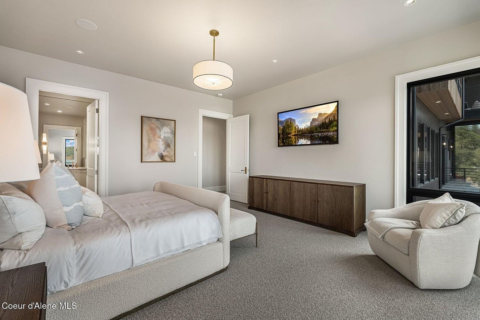 This is a primary bedroom featuring a large bed with a textured base and light-colored bedding. A modern drum pendant light hangs from the ceiling, and a long wooden cabinet sits beneath a wall-mounted television. The room also includes a comfortable armchair and a large window offering natural light.