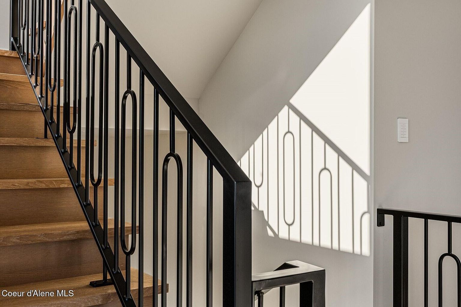 This image showcases a modern staircase with wooden steps and a sleek, black metal railing featuring a unique, rounded design. Sunlight streams in, casting interesting shadows on the wall and highlighting the clean lines of the architectural details. The overall impression is one of contemporary elegance and bright, airy design.