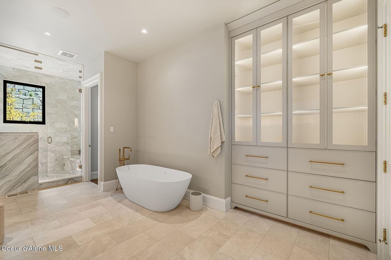 This is a primary bathroom featuring a freestanding white bathtub with gold fixtures, a glass-enclosed shower with marble accents, and a built-in storage unit with glass doors and gold hardware. The flooring is light-colored tile, and the walls are a neutral tone, creating a serene and luxurious atmosphere. The perspective is from the doorway, showcasing the layout and high-end finishes.