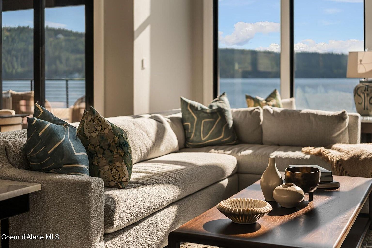 This is an interior shot of a living room featuring a large, comfortable-looking sectional sofa adorned with decorative pillows. A dark wood coffee table displays various decorative bowls and vases. Large windows offer a scenic view of a lake and surrounding landscape, creating a bright and inviting atmosphere.