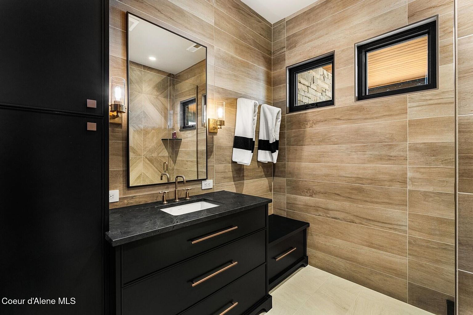 This is a modern bathroom featuring wood-look tile walls and a dark vanity with copper hardware. A large mirror hangs above the sink, flanked by sconce lighting. Two small windows provide natural light, and towels hang neatly on a rack, creating a clean and stylish space.