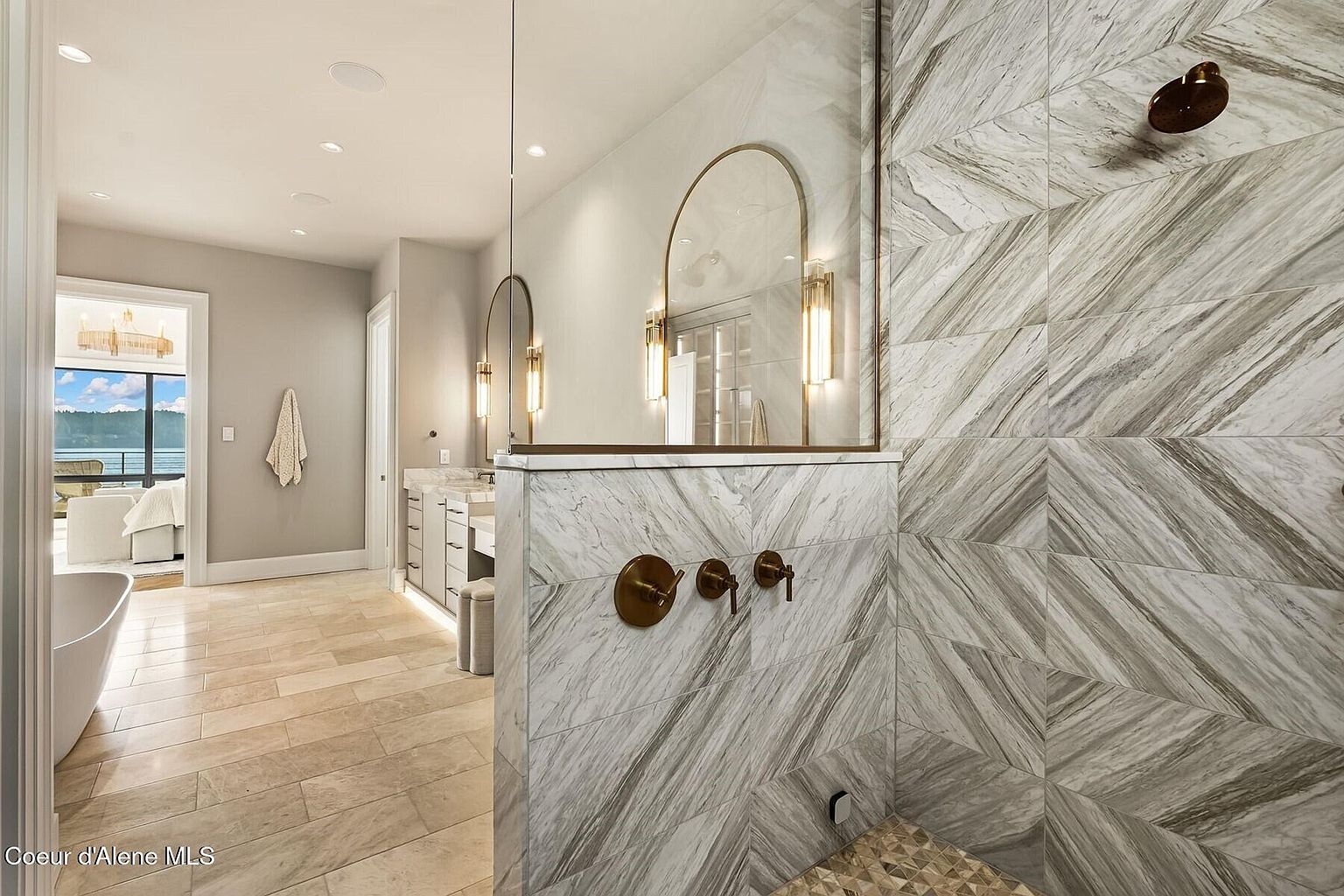 This is a luxurious primary bathroom featuring a modern shower with marble-like tile in a chevron pattern and bronze fixtures. The bathroom includes a vanity area with arched mirrors and pendant lighting. The flooring is light-colored tile, and there's a glimpse of a freestanding tub and a view of a lake through a doorway, creating a spa-like atmosphere.