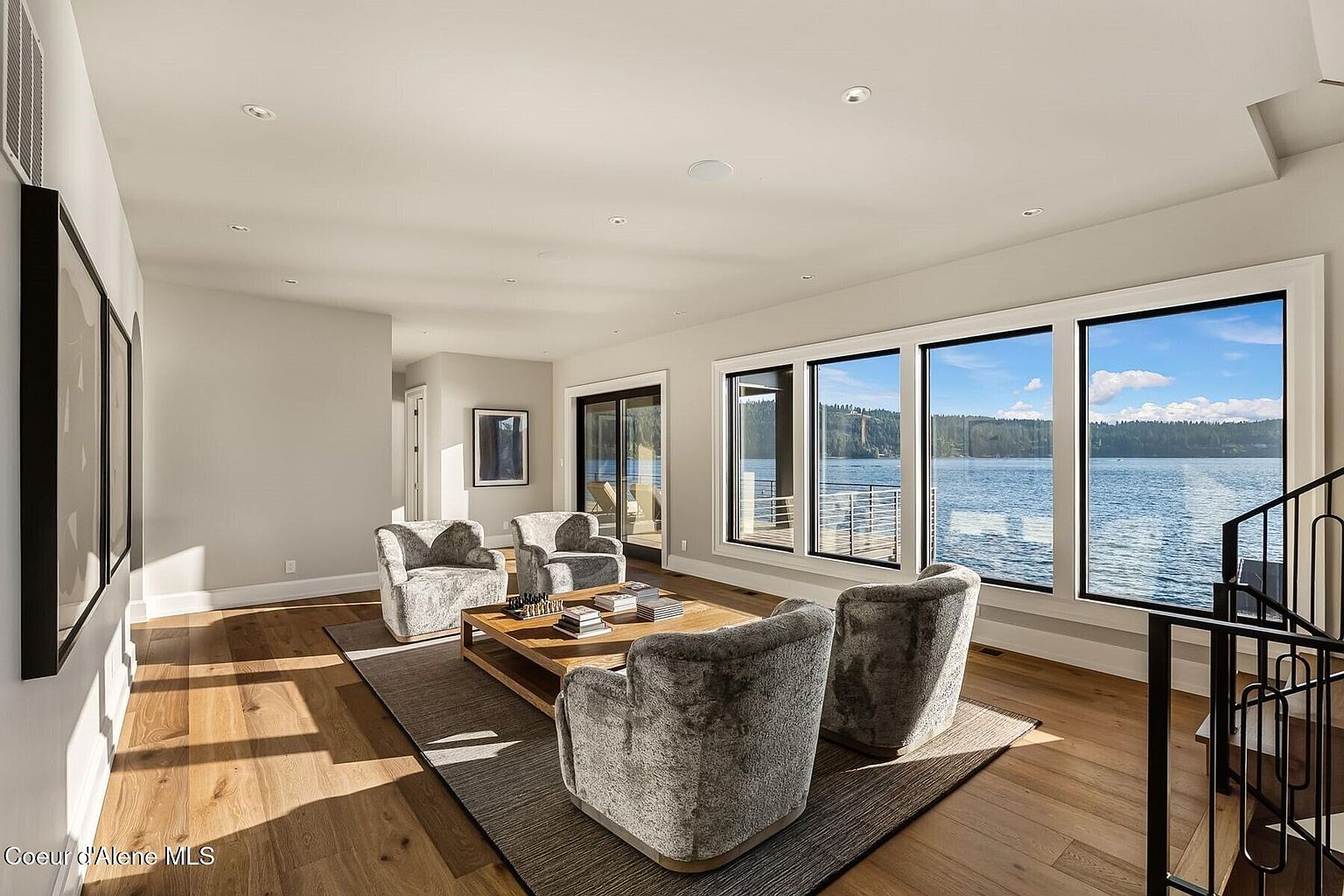This is an interior shot of a living room featuring four plush armchairs arranged around a wooden coffee table on a dark rug. Large windows offer a view of a lake, bringing natural light into the space. The room has hardwood floors and a neutral color palette, creating a serene and inviting atmosphere.
