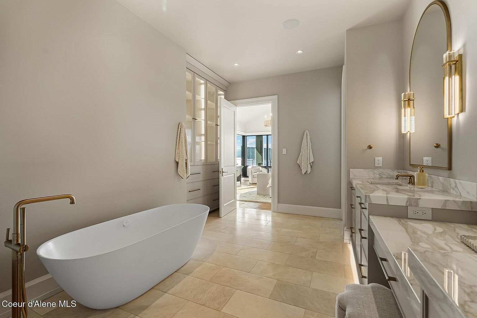 This is a luxurious primary bathroom featuring a freestanding white bathtub with a gold faucet. The room has beige tile flooring and light gray walls. A double vanity with marble countertops and a large arched mirror with sconces adds to the elegant aesthetic. A doorway leads to another room with a view of the outside.