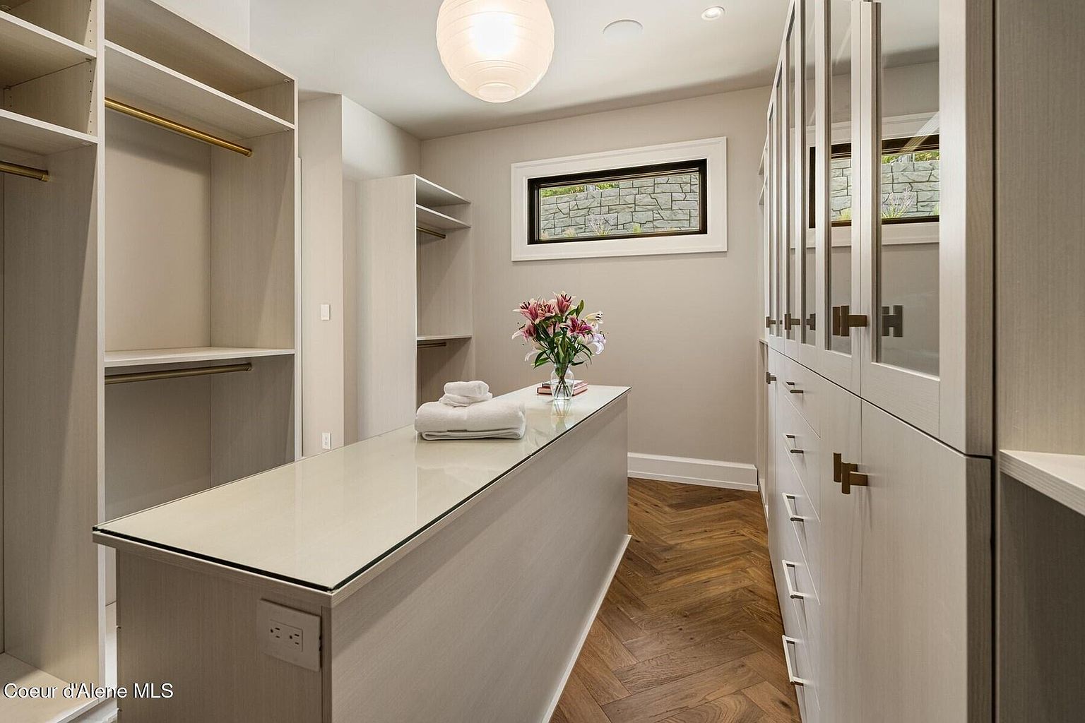 This is a well-organized walk-in closet featuring custom shelving and cabinetry in a light neutral tone. A central island with a light countertop provides additional storage and display space, topped with fresh flowers and neatly folded towels. The herringbone wood flooring adds a touch of elegance, and the overall impression is one of luxury and functionality.