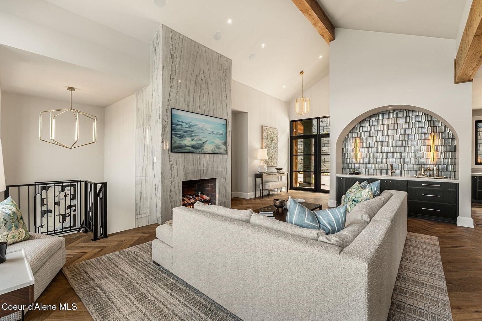 This is an interior shot of a modern living room featuring a marble fireplace with a mounted television. The room is decorated with a neutral-toned sectional sofa, patterned throw pillows, and a textured area rug. The hardwood flooring is laid in a herringbone pattern, and the room has high ceilings with exposed wooden beams, creating a spacious and luxurious atmosphere.