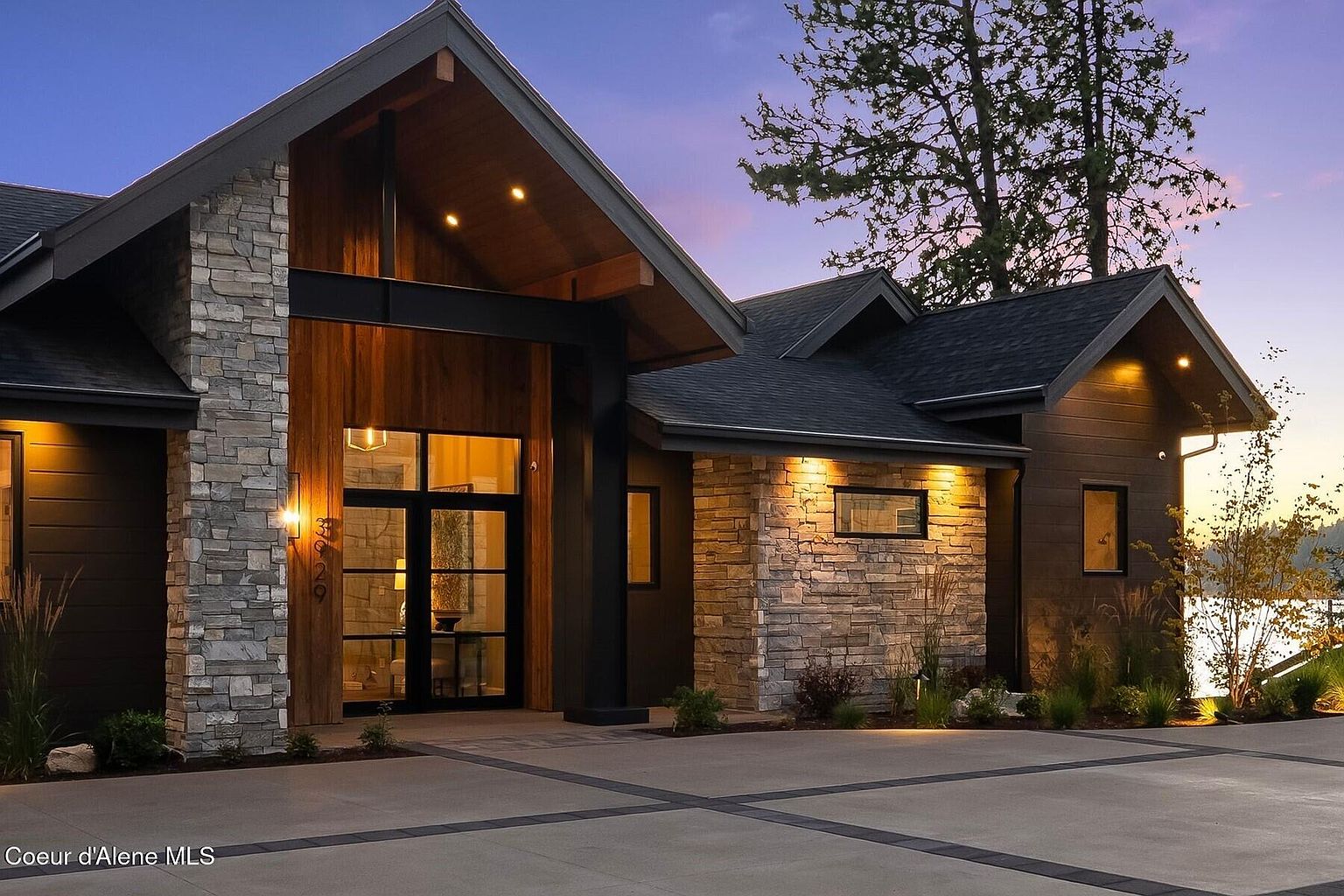 This is a front exterior view of a modern home at dusk, showcasing a blend of stone, wood, and dark metal accents. The architectural design features a prominent gabled entrance with warm interior lighting visible through the glass front door. The well-lit driveway and landscaping enhance the property's curb appeal, creating an inviting and sophisticated impression.