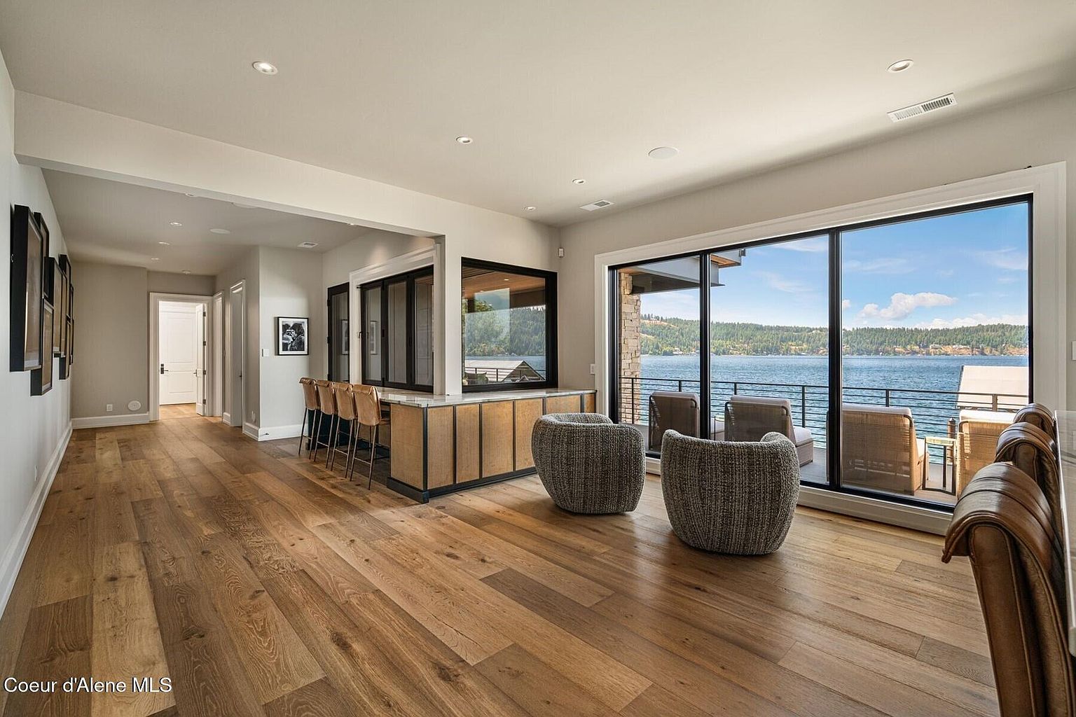This is a spacious living room featuring hardwood floors and a bar area with seating. Large windows offer a stunning view of the lake and surrounding landscape, bringing natural light into the room. The room is furnished with modern chairs and bar stools, creating a comfortable and stylish space for relaxation and entertainment.
