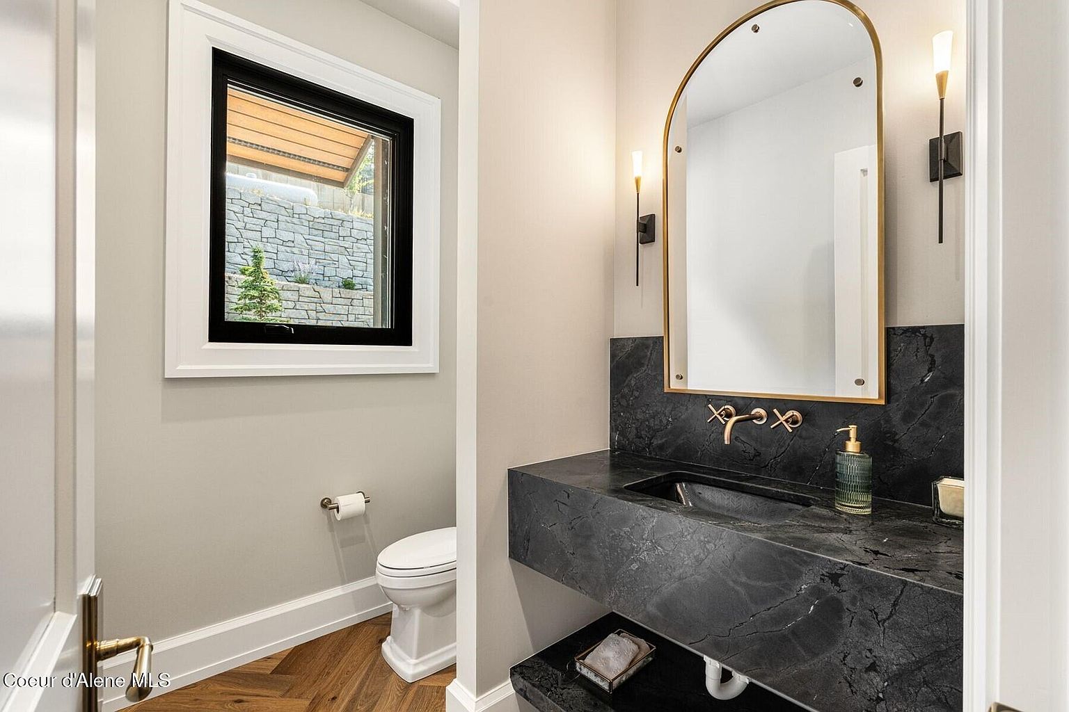 This is a stylish powder room featuring a black marble countertop and backsplash with a vessel sink and gold fixtures. An arched mirror with a gold frame is mounted above the sink, flanked by sconce lighting. A window provides natural light, and the herringbone wood flooring adds a touch of elegance.