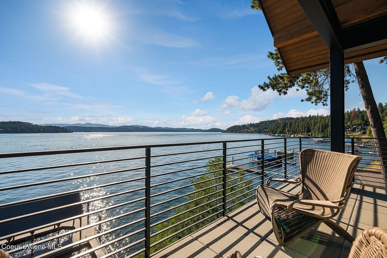 This image showcases a stunning balcony view overlooking a serene lake, framed by a modern railing. A comfortable wicker chair invites relaxation, while the sun glistens on the water, creating a tranquil atmosphere. The scene emphasizes the property's desirable waterfront location and outdoor living space.