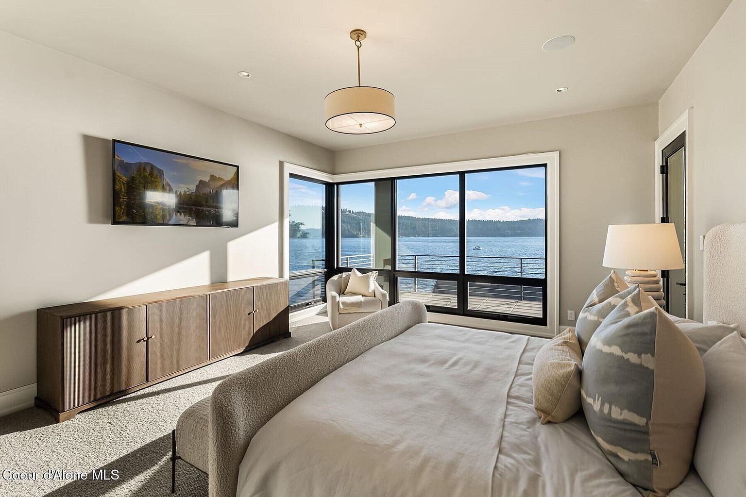 This is a primary bedroom featuring a large bed with neutral bedding and decorative pillows, a modern dresser with a TV above it, and a sitting area by a large window with a water view. The room has a contemporary style with neutral color palette and recessed lighting, creating a serene and luxurious atmosphere.
