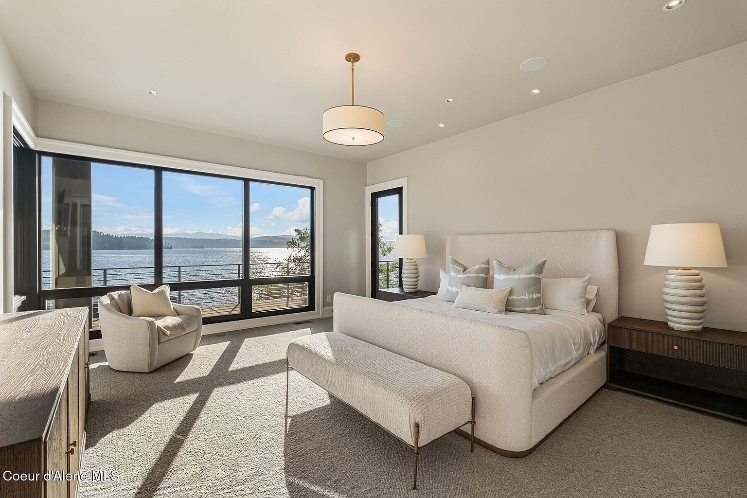 This is a bright and airy primary bedroom with a large window offering a scenic lake view. The room features a neutral color palette, a plush upholstered bed with a bench, and stylish bedside tables with lamps. The overall impression is one of luxury and tranquility.