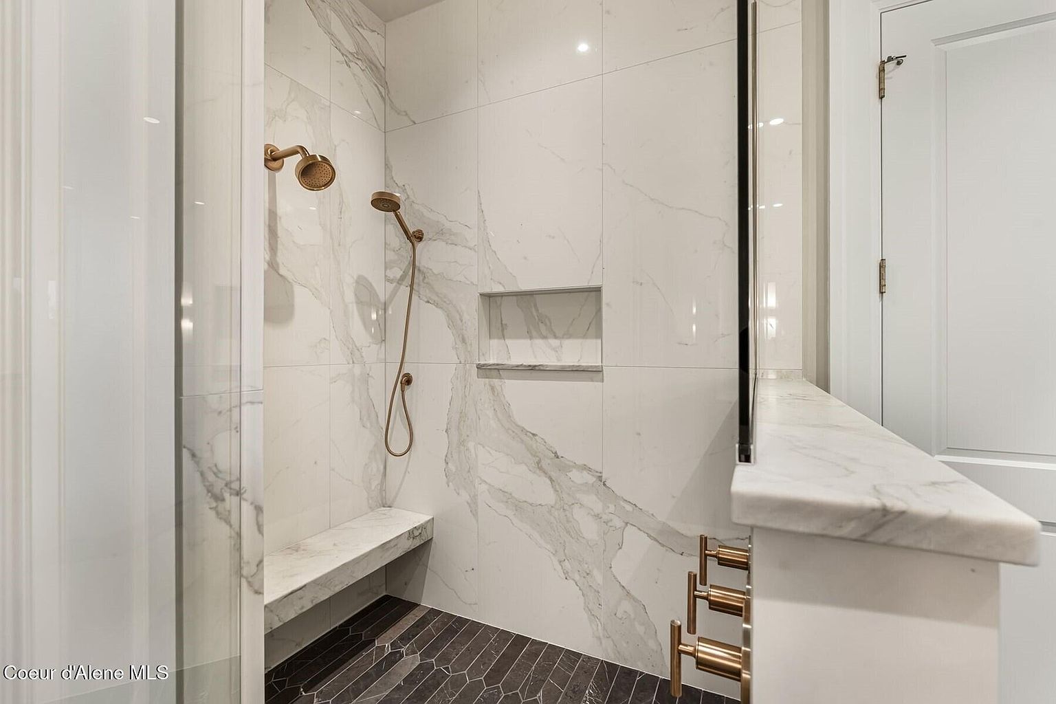 This is a luxurious primary bathroom featuring a walk-in shower with marble-like tile walls, a built-in bench, and dual shower heads in a bronze finish. The shower also includes a recessed niche for storage. The floor is tiled in a dark, geometric pattern, and a heated towel rack adds a touch of comfort.