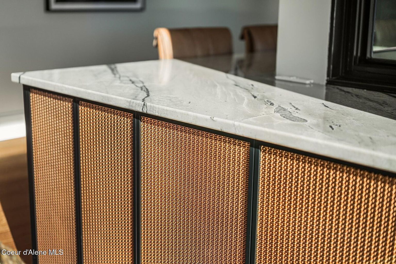The image showcases a detailed view of a kitchen island or counter featuring a light marble countertop with dark veining. The base of the island is adorned with a woven, possibly copper or bronze-toned, material framed by dark trim, adding a touch of texture and sophistication. The overall impression is one of high-end design and attention to detail.