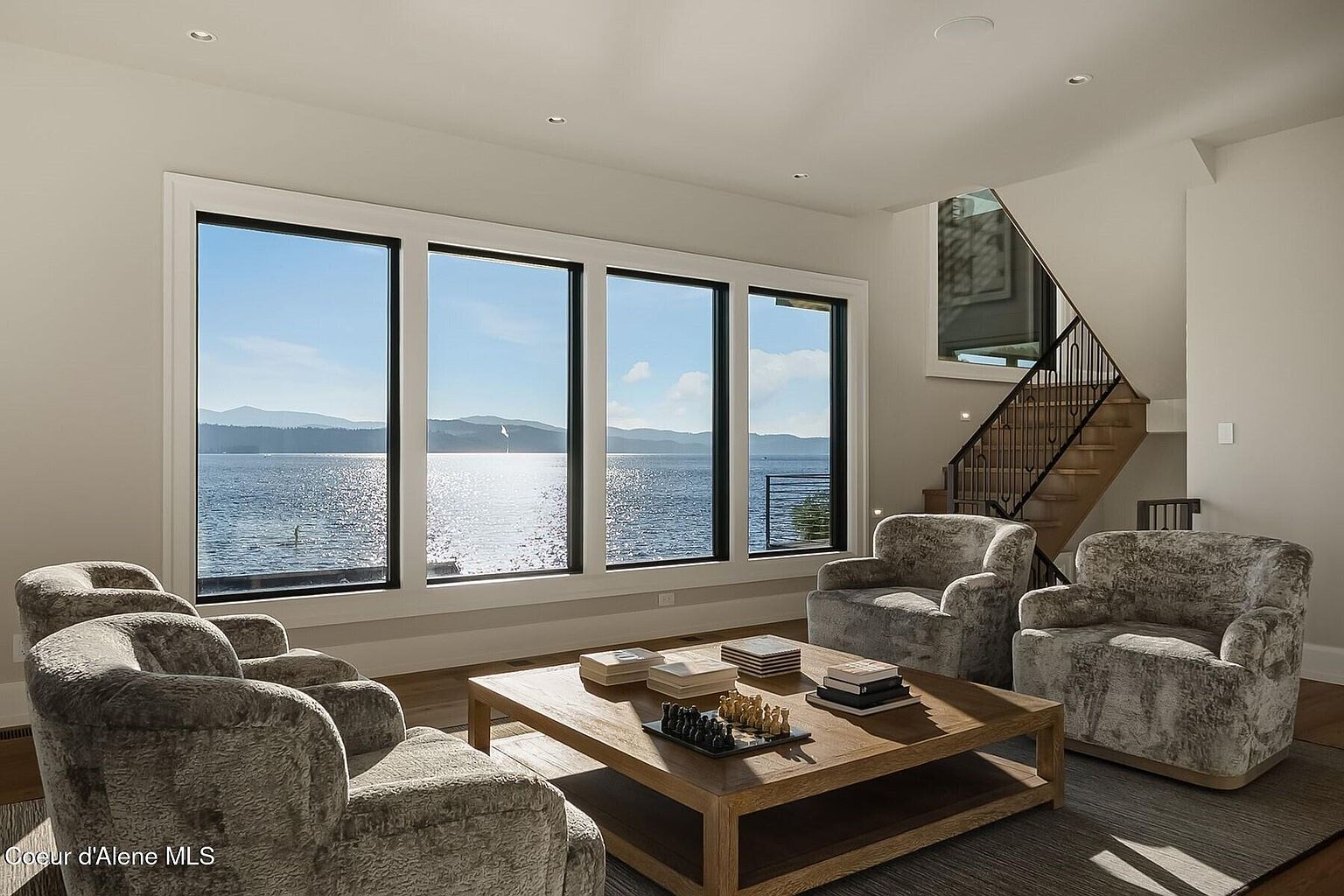 This is an interior shot of a living room featuring a stunning lake view through large windows. The room is furnished with comfortable armchairs arranged around a wooden coffee table, creating a cozy and inviting atmosphere. A staircase is visible in the background, adding architectural interest to the space.