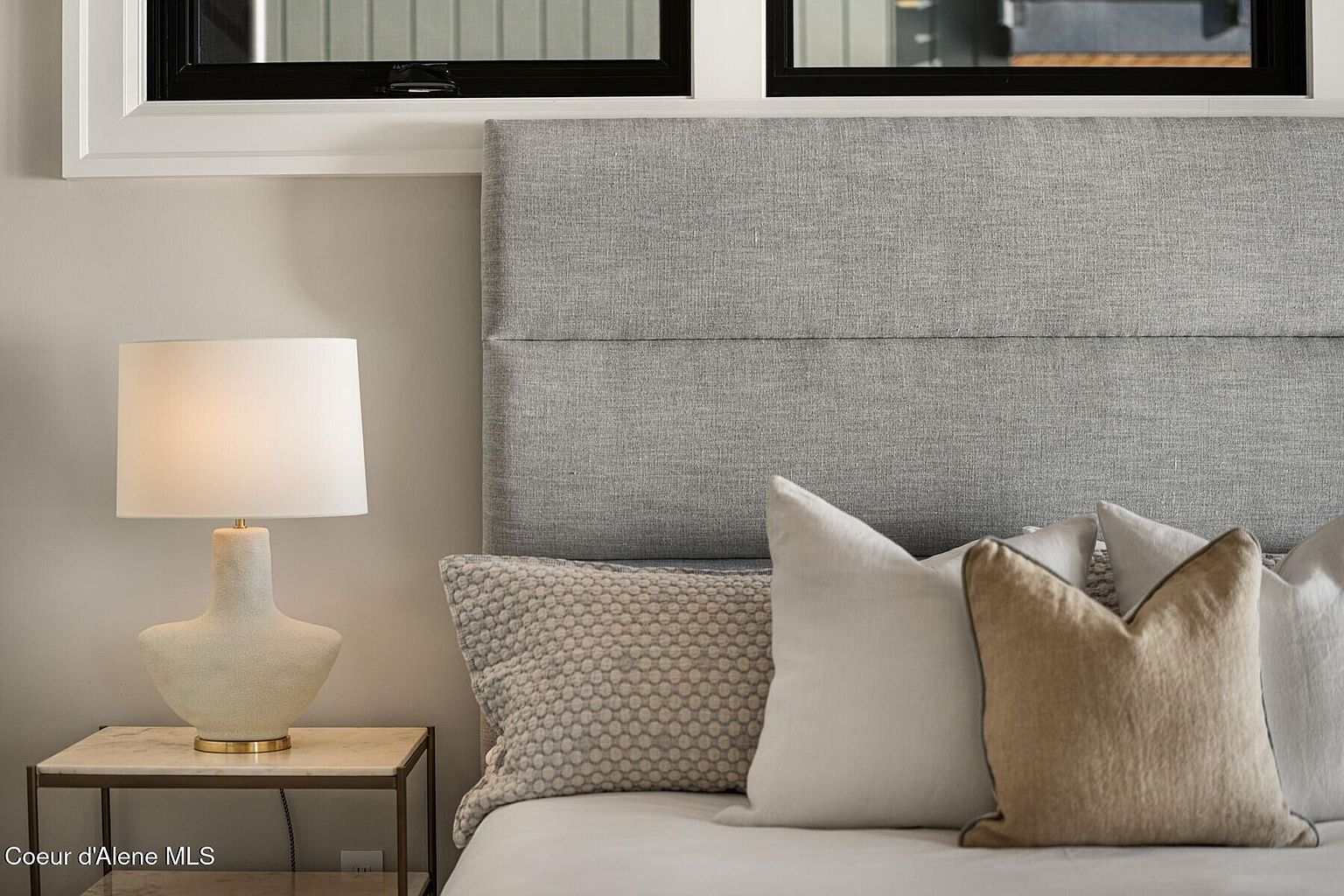 This is a close-up shot of a bedroom featuring a gray upholstered headboard, several decorative pillows in neutral tones, and a bedside table with a modern lamp. The color palette is soft and calming, creating a serene and inviting atmosphere. The perspective is from the side of the bed, focusing on the headboard and pillows.