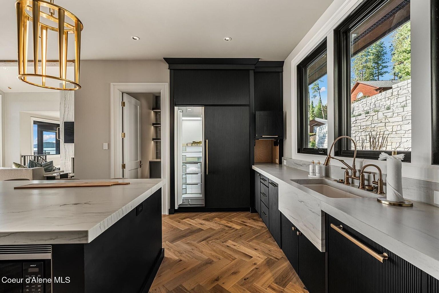This is a modern kitchen featuring black cabinetry, a marble countertop, and herringbone wood floors. The kitchen includes a large island, a built-in refrigerator, and a sink with gold fixtures. The space is well-lit with natural light from the windows and a statement gold chandelier, creating a luxurious and inviting atmosphere.
