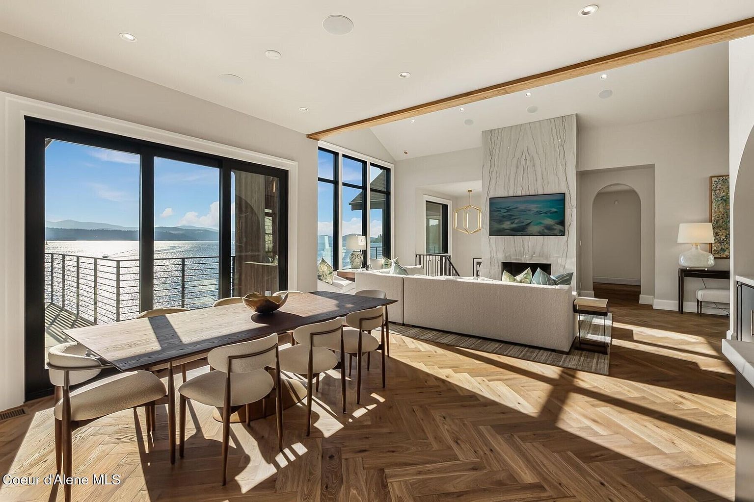 This interior shot showcases a dining area seamlessly integrated with a living space, featuring a long wooden dining table with modern chairs. Large sliding glass doors offer stunning waterfront views, enhancing the room's bright and airy atmosphere. The herringbone wood flooring adds a touch of elegance, while the open layout promotes a sense of spaciousness and connectivity.